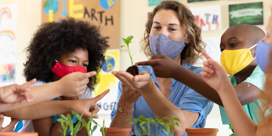 Imagen de una maestra y estudiantes con mascarillas y que miran los plantine