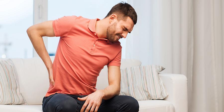 Hombre sentado en el sofá sujetándose la parte baja de la espalda mientras hace muecas de dolor.