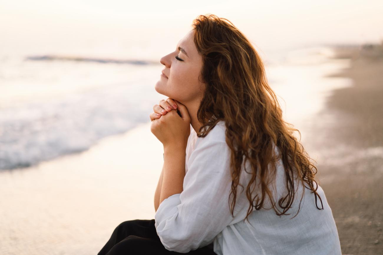 Imagen de una mujer con los ojos cerrados y las manos entrelazadas sentada junto al mar.