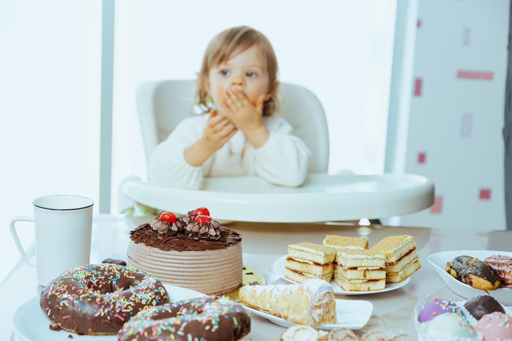 Imagen de una niña pequeña sentada en una silla alta enfrente de una variedad de tortas y postres ubicados sobre una mesa.