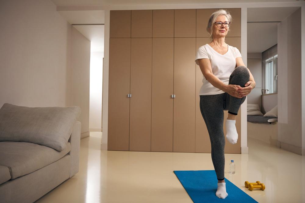 Imagen de una mujer mayor parada en una pierna sobre una colchoneta de yoga, mientras sujeta con sus manos su otra pierna.