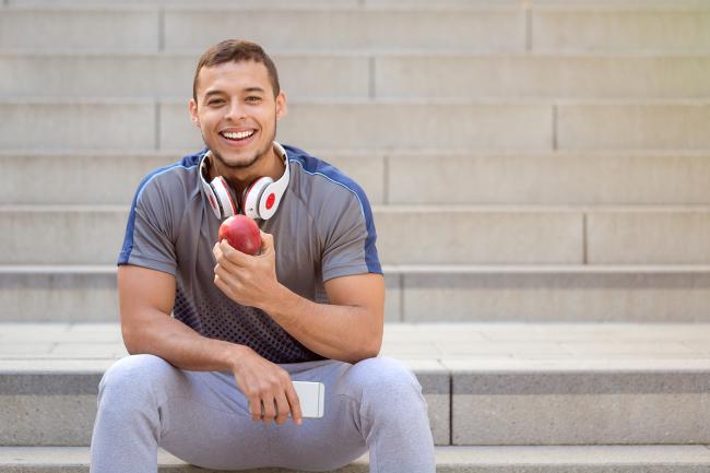 Un joven sonriente vestido con ropa deportiva sentado en un escalón comiendo una manzana
