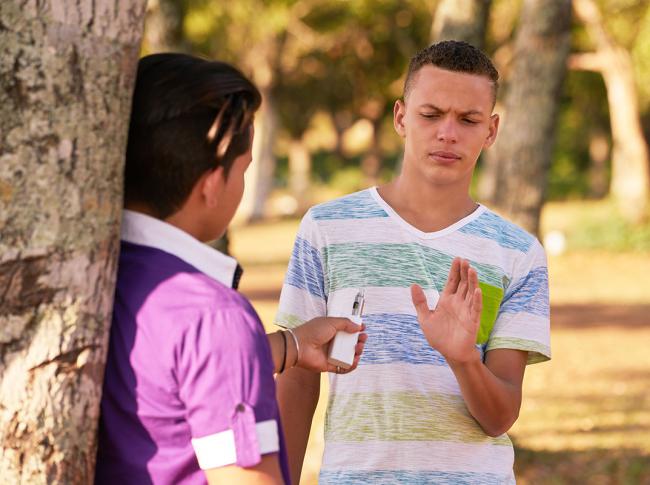 Fotografía de un adolescente con su rostro preocupado rechazando un vaporizador de tabaco que le ofrece otro joven. Ambos están en un parque.