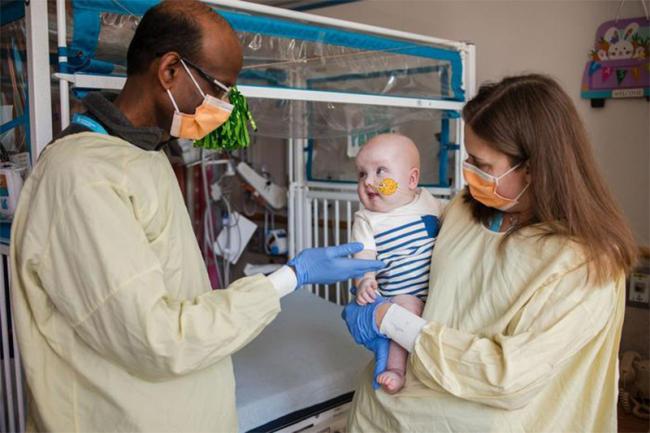 Una fotografía muestra a una doctora con un bebé en sus brazos, paciente de un tratamiento genético personalizado para tratar un trastorno potencialmente mortal. Otro doctor está saludando al bebé. Los doctores están usando mascarillas.