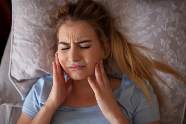 Una fotografía muestra a una mujer tocando su mandíbula con sus manos y apretando sus dientes mientras está recostada. Su rostro tiene una expresión de dolor.