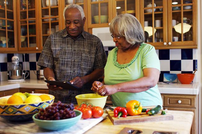 Una fotografía muestra a una pareja de adultos mayores preparando juntos alimentos saludables en la cocina. Están sonriendo.