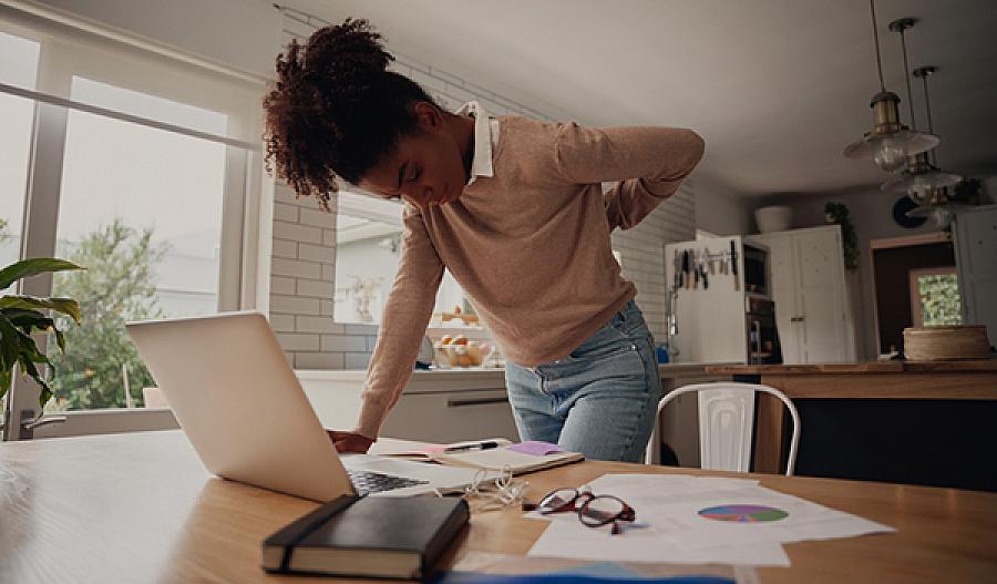 Imagen de una mujer joven de pie y sujetándose la espalda mientras trabaja en una laptop en casa
