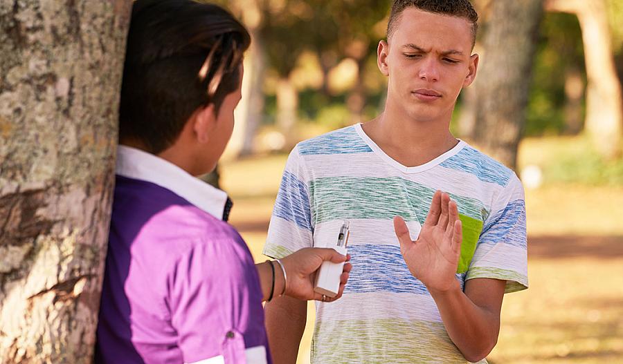 Fotografía de un adolescente con su rostro preocupado rechazando un vaporizador de tabaco que le ofrece otro joven. Ambos están en un parque.