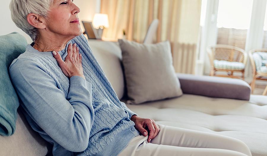 Fotografía de una adulta mayor está sentada en un sofá, mientras coloca su mano en su pecho. Su rostro muestra dolor.