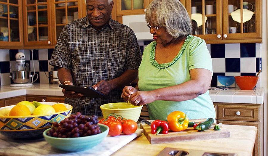Una fotografía muestra a una pareja de adultos mayores preparando juntos alimentos saludables en la cocina. Están sonriendo.
