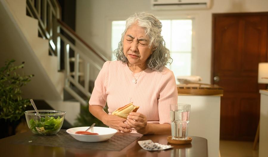 Una fotografía muestra a una adulta mayor comiendo un sándwich, al tiempo que cierra sus ojos en actitud de dolor, mientras toca su estómago con una de sus manos.