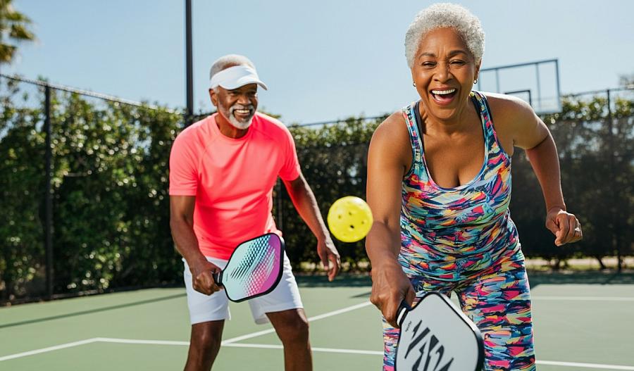 Una fotografía muestra a dos adultos mayores, un hombre y una mujer, jugando pickleball. Están riéndose, en un día soleado.